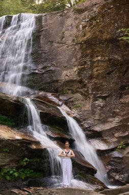 Güzel genç bir kadın gün batımında şelale ve nehir kenarında ağır ağır tai chi hareketleri yapıyor. Doğadaki uyum dengesini ve uygunluğu yakalıyor.