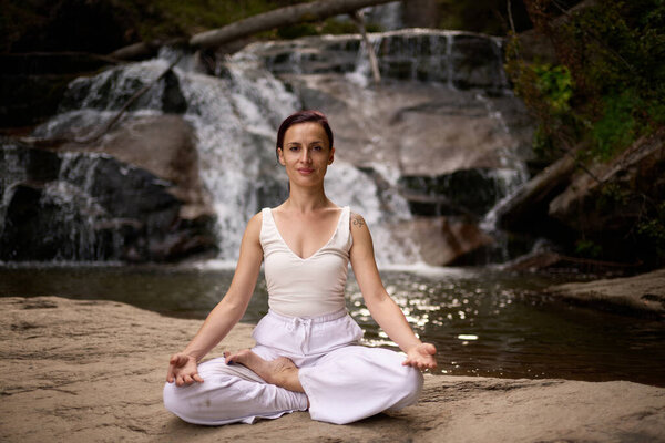 Young woman sitting in lotus pose under tropical waterfall meditating with calm strength and mindfulness fully connected to nature energy tranquility and inner balance