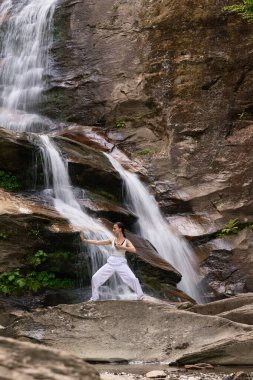 Güzel genç bir kadın gün batımında şelale ve nehir kenarında ağır ağır tai chi hareketleri yapıyor. Doğadaki uyum dengesini ve uygunluğu yakalıyor.