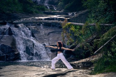 Yoga yapan kadın, gün doğumunda şelalenin yanında poz veriyor. Açık havada sağlıklı meditasyon yapıyor. Rahatlama farkındalık ahenk ruhsal geri çekilme sükunet konsepti.