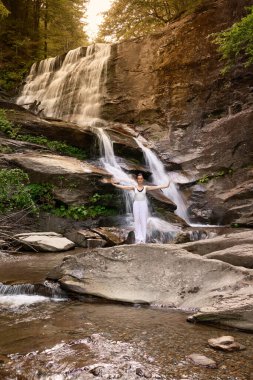 Güzel genç bir kadın gün batımında şelale ve nehir kenarında ağır ağır tai chi hareketleri yapıyor. Doğadaki uyum dengesini ve uygunluğu yakalıyor.
