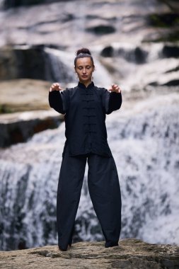 Doğada Tai Chi uygulayan, geleneksel bir siyah dövüş sanatları üniforması giyen, bir şelalenin yanında zarif bir şekilde duran, meditasyon ve dengeye odaklanmış bir kadın. Asya kültüründe sağlıklı yaşam tarzı 