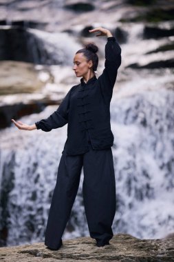 Doğada Tai Chi uygulayan, geleneksel bir siyah dövüş sanatları üniforması giyen, bir şelalenin yanında zarif bir şekilde duran, meditasyon ve dengeye odaklanmış bir kadın. Asya kültüründe sağlıklı yaşam tarzı 