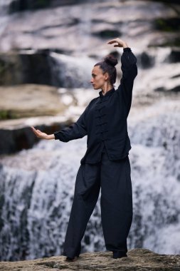 Doğada Tai Chi uygulayan, geleneksel bir siyah dövüş sanatları üniforması giyen, bir şelalenin yanında zarif bir şekilde duran, meditasyon ve dengeye odaklanmış bir kadın. Asya kültüründe sağlıklı yaşam tarzı 
