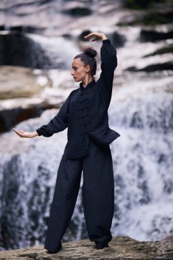 Doğada Tai Chi uygulayan, geleneksel bir siyah dövüş sanatları üniforması giyen, bir şelalenin yanında zarif bir şekilde duran, meditasyon ve dengeye odaklanmış bir kadın. Asya kültüründe sağlıklı yaşam tarzı 