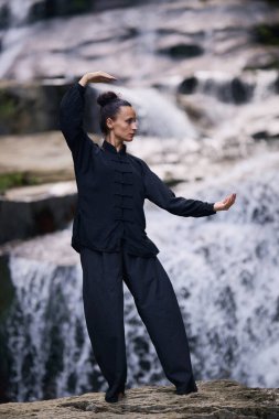 Doğada Tai Chi uygulayan, geleneksel bir siyah dövüş sanatları üniforması giyen, bir şelalenin yanında zarif bir şekilde duran, meditasyon ve dengeye odaklanmış bir kadın. Asya kültüründe sağlıklı yaşam tarzı 