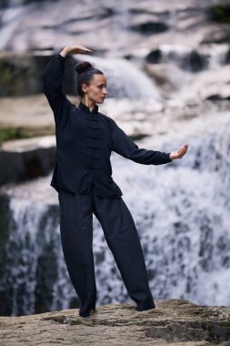 Doğada Tai Chi uygulayan, geleneksel bir siyah dövüş sanatları üniforması giyen, bir şelalenin yanında zarif bir şekilde duran, meditasyon ve dengeye odaklanmış bir kadın. Asya kültüründe sağlıklı yaşam tarzı 