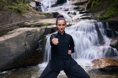 Doğada Tai Chi uygulayan, geleneksel bir siyah dövüş sanatları üniforması giyen, bir şelalenin yanında zarif bir şekilde duran, meditasyon ve dengeye odaklanmış bir kadın. Asya kültüründe sağlıklı yaşam tarzı 