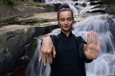 Doğada Tai Chi uygulayan, geleneksel bir siyah dövüş sanatları üniforması giyen, bir şelalenin yanında zarif bir şekilde duran, meditasyon ve dengeye odaklanmış bir kadın. Asya kültüründe sağlıklı yaşam tarzı 
