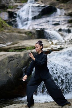 Güzel genç bir kadın gün batımında şelale ve nehir kenarında ağır ağır tai chi hareketleri yapıyor. Doğadaki uyum dengesini ve uygunluğu yakalıyor..