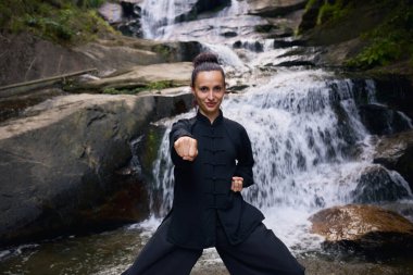 Doğada Tai Chi uygulayan, geleneksel bir siyah dövüş sanatları üniforması giyen, bir şelalenin yanında zarif bir şekilde duran, meditasyon ve dengeye odaklanmış bir kadın. Asya kültüründe sağlıklı yaşam tarzı 