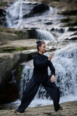 Güzel genç bir kadın gün batımında şelale ve nehir kenarında ağır ağır tai chi hareketleri yapıyor. Doğadaki uyum dengesini ve uygunluğu yakalıyor..