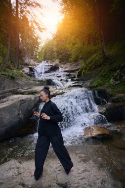 Doğada Tai Chi uygulayan, geleneksel bir siyah dövüş sanatları üniforması giyen, bir şelalenin yanında zarif bir şekilde duran, meditasyon ve dengeye odaklanmış bir kadın. Asya kültüründe sağlıklı yaşam tarzı 