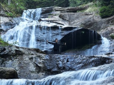 Doğada Tai Chi uygulayan, geleneksel bir siyah dövüş sanatları üniforması giyen, bir şelalenin yanında zarif bir şekilde duran, meditasyon ve dengeye odaklanmış bir kadın. Asya kültüründe sağlıklı yaşam tarzı 