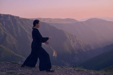 Güneşin doğuşunda bir dağda geleneksel siyah bir kıyafetle Tai Chi çalışması yapan bir kadın, kollarını meditasyon pozunda uzatarak, doğada denge, enerji ve farkındalık üzerine odaklanır.. 