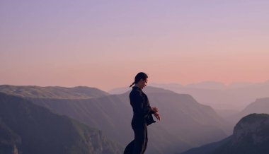 Güneşin doğuşunda bir dağda geleneksel siyah bir kıyafetle Tai Chi çalışması yapan bir kadın, kollarını meditasyon pozunda uzatarak, doğada denge, enerji ve farkındalık üzerine odaklanır.. 