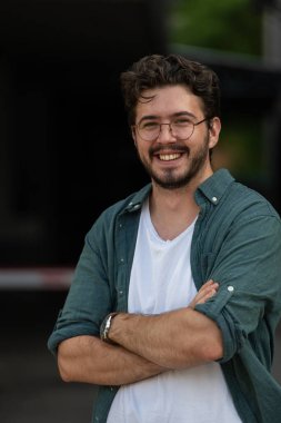 Tişörtlü başarılı genç bir iş adamı, kollarını kavuşturmuş, açık havada poz veriyor, yüzünde kendinden emin bir ifade var.