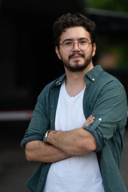 Tişörtlü başarılı genç bir iş adamı, kollarını kavuşturmuş, açık havada poz veriyor, yüzünde kendinden emin bir ifade var.