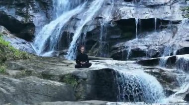 Yavaş tai chi hareketleri yapan ya da şelalenin yanında yoga yapan bir kadının hava aracı görüntüsü. Kolları açmak, ahengi ifade etmek, lotus pozisyonunda oturmak, farkındalık, zindelik ve ruhanilik.