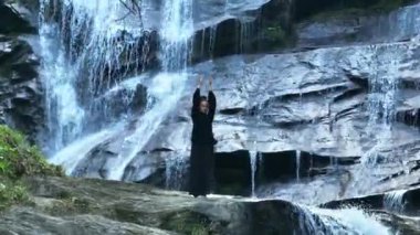 Yavaş tai chi hareketleri yapan ya da şelalenin yanında yoga yapan bir kadının hava aracı görüntüsü. Kolları açmak, ahengi ifade etmek, lotus pozisyonunda oturmak, farkındalık, zindelik ve ruhanilik.