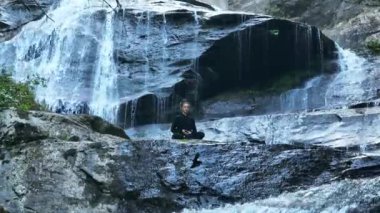 Yavaş tai chi hareketleri yapan ya da şelalenin yanında yoga yapan bir kadının hava aracı görüntüsü. Kolları açmak, ahengi ifade etmek, lotus pozisyonunda oturmak, farkındalık, zindelik ve ruhanilik.