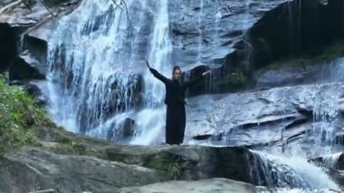 Yavaş tai chi hareketleri yapan ya da şelalenin yanında yoga yapan bir kadının hava aracı görüntüsü. Kolları açmak, ahengi ifade etmek, lotus pozisyonunda oturmak, farkındalık, zindelik ve ruhanilik.