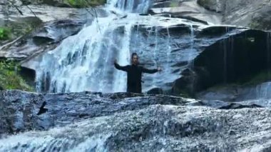 Yavaş tai chi hareketleri yapan ya da şelalenin yanında yoga yapan bir kadının hava aracı görüntüsü. Kolları açmak, ahengi ifade etmek, lotus pozisyonunda oturmak, farkındalık, zindelik ve ruhanilik.