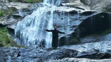 Yavaş tai chi hareketleri yapan ya da şelalenin yanında yoga yapan bir kadının hava aracı görüntüsü. Kolları açmak, ahengi ifade etmek, lotus pozisyonunda oturmak, farkındalık, zindelik ve ruhanilik.