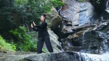Yavaş tai chi hareketleri yapan ya da şelalenin yanında yoga yapan bir kadının hava aracı görüntüsü. Kolları açmak, ahengi ifade etmek, lotus pozisyonunda oturmak, farkındalık, zindelik ve ruhanilik.