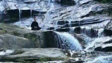 Yavaş tai chi hareketleri yapan ya da şelalenin yanında yoga yapan bir kadının hava aracı görüntüsü. Kolları açmak, ahengi ifade etmek, lotus pozisyonunda oturmak, farkındalık, zindelik ve ruhanilik.