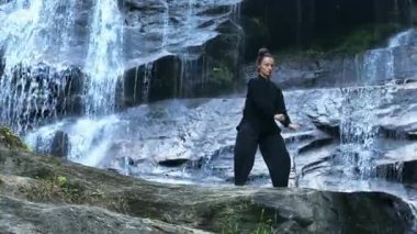 Yavaş tai chi hareketleri yapan ya da şelalenin yanında yoga yapan bir kadının hava aracı görüntüsü. Kolları açmak, ahengi ifade etmek, lotus pozisyonunda oturmak, farkındalık, zindelik ve ruhanilik.