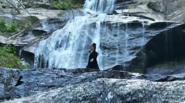 Yavaş tai chi hareketleri yapan ya da şelalenin yanında yoga yapan bir kadının hava aracı görüntüsü. Kolları açmak, ahengi ifade etmek, lotus pozisyonunda oturmak, farkındalık, zindelik ve ruhanilik.