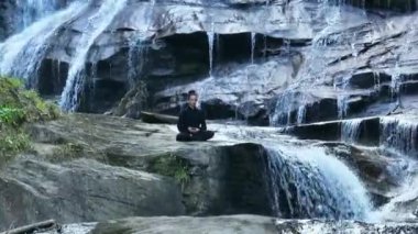 Yavaş tai chi hareketleri yapan ya da şelalenin yanında yoga yapan bir kadının hava aracı görüntüsü. Kolları açmak, ahengi ifade etmek, lotus pozisyonunda oturmak, farkındalık, zindelik ve ruhanilik.