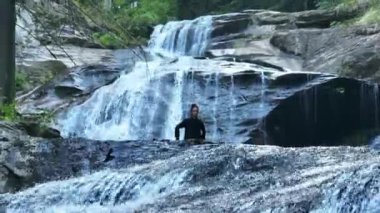 Yavaş tai chi hareketleri yapan ya da şelalenin yanında yoga yapan bir kadının hava aracı görüntüsü. Kolları açmak, ahengi ifade etmek, lotus pozisyonunda oturmak, farkındalık, zindelik ve ruhanilik.