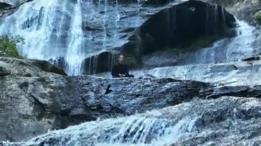Yavaş tai chi hareketleri yapan ya da şelalenin yanında yoga yapan bir kadının hava aracı görüntüsü. Kolları açmak, ahengi ifade etmek, lotus pozisyonunda oturmak, farkındalık, zindelik ve ruhanilik.