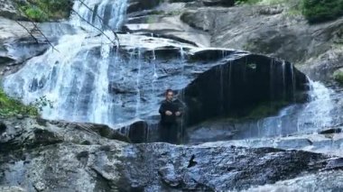 Yavaş tai chi hareketleri yapan ya da şelalenin yanında yoga yapan bir kadının hava aracı görüntüsü. Kolları açmak, ahengi ifade etmek, lotus pozisyonunda oturmak, farkındalık, zindelik ve ruhanilik.