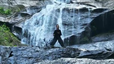 Yavaş tai chi hareketleri yapan ya da şelalenin yanında yoga yapan bir kadının hava aracı görüntüsü. Kolları açmak, ahengi ifade etmek, lotus pozisyonunda oturmak, farkındalık, zindelik ve ruhanilik.