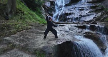 Yavaş tai chi hareketleri yapan ya da şelalenin yanında yoga yapan bir kadının hava aracı görüntüsü. Kolları açmak, ahengi ifade etmek, lotus pozisyonunda oturmak, farkındalık, zindelik ve ruhanilik.