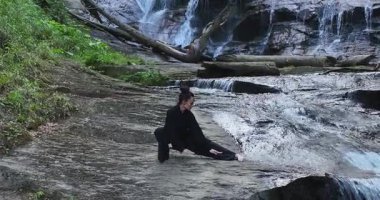 Yavaş tai chi hareketleri yapan ya da şelalenin yanında yoga yapan bir kadının hava aracı görüntüsü. Kolları açmak, ahengi ifade etmek, lotus pozisyonunda oturmak, farkındalık, zindelik ve ruhanilik.