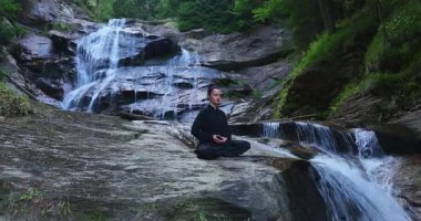 Yavaş tai chi hareketleri yapan ya da şelalenin yanında yoga yapan bir kadının hava aracı görüntüsü. Kolları açmak, ahengi ifade etmek, lotus pozisyonunda oturmak, farkındalık, zindelik ve ruhanilik.