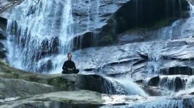 Yavaş tai chi hareketleri yapan ya da şelalenin yanında yoga yapan bir kadının hava aracı görüntüsü. Kolları açmak, ahengi ifade etmek, lotus pozisyonunda oturmak, farkındalık, zindelik ve ruhanilik.