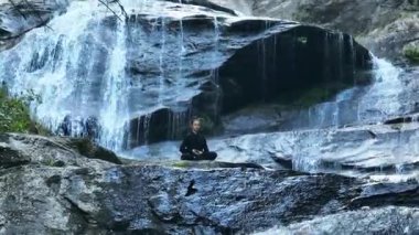 Yavaş tai chi hareketleri yapan ya da şelalenin yanında yoga yapan bir kadının hava aracı görüntüsü. Kolları açmak, ahengi ifade etmek, lotus pozisyonunda oturmak, farkındalık, zindelik ve ruhanilik.