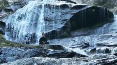 Yavaş tai chi hareketleri yapan ya da şelalenin yanında yoga yapan bir kadının hava aracı görüntüsü. Kolları açmak, ahengi ifade etmek, lotus pozisyonunda oturmak, farkındalık, zindelik ve ruhanilik.