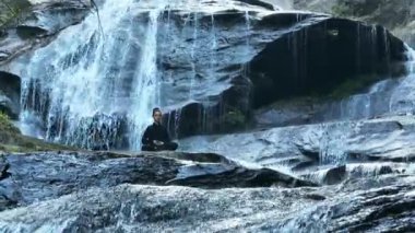 Yavaş tai chi hareketleri yapan ya da şelalenin yanında yoga yapan bir kadının hava aracı görüntüsü. Kolları açmak, ahengi ifade etmek, lotus pozisyonunda oturmak, farkındalık, zindelik ve ruhanilik.