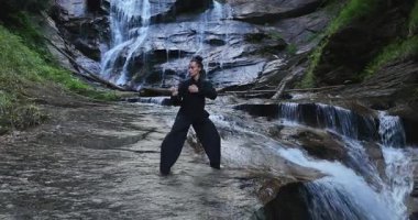 Yavaş tai chi hareketleri yapan ya da şelalenin yanında yoga yapan bir kadının hava aracı görüntüsü. Kolları açmak, ahengi ifade etmek, lotus pozisyonunda oturmak, farkındalık, zindelik ve ruhanilik.