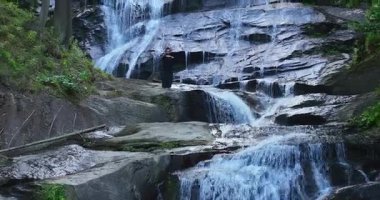 Yavaş tai chi hareketleri yapan ya da şelalenin yanında yoga yapan bir kadının hava aracı görüntüsü. Kolları açmak, ahengi ifade etmek, lotus pozisyonunda oturmak, farkındalık, zindelik ve ruhanilik.