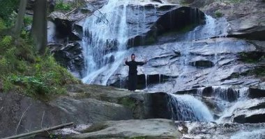 Yavaş tai chi hareketleri yapan ya da şelalenin yanında yoga yapan bir kadının hava aracı görüntüsü. Kolları açmak, ahengi ifade etmek, lotus pozisyonunda oturmak, farkındalık, zindelik ve ruhanilik.