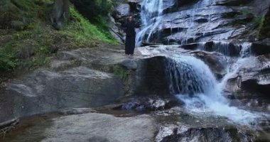 Yavaş tai chi hareketleri yapan ya da şelalenin yanında yoga yapan bir kadının hava aracı görüntüsü. Kolları açmak, ahengi ifade etmek, lotus pozisyonunda oturmak, farkındalık, zindelik ve ruhanilik.