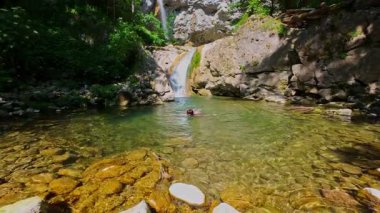 Güneşli bir günde yemyeşil orman ve kayalarla çevrili temiz bir dağ nehrinde yüzen bir kadının FPV drone görüntüsü. Vahşi doğada yaz kaçışı, macera ve seyahat kavramının tazelenmesi 
