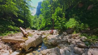 Güneşli bir günde yemyeşil orman ve kayalarla çevrili temiz bir dağ nehrinde yüzen bir kadının FPV drone görüntüsü. Vahşi doğada yaz kaçışı, macera ve seyahat kavramının tazelenmesi 